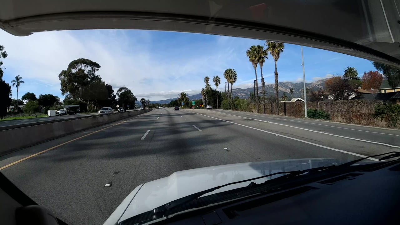 Highway lanes with palm trees, low roadside buildings, and hills in the distance.
