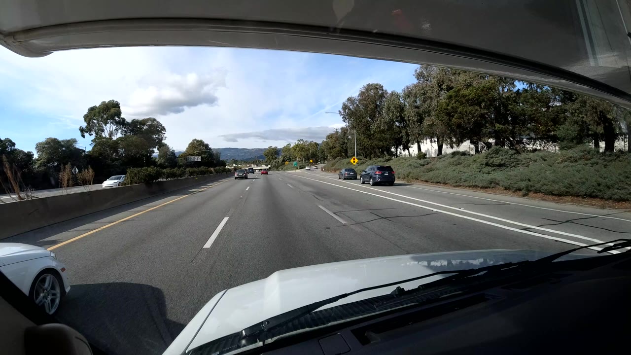 A Coast California highway is seen through a vehicle windshield at the start of the clip, with the white hood in the foreground, cars ahead, trees along both sides, distant hills, and no visible people.