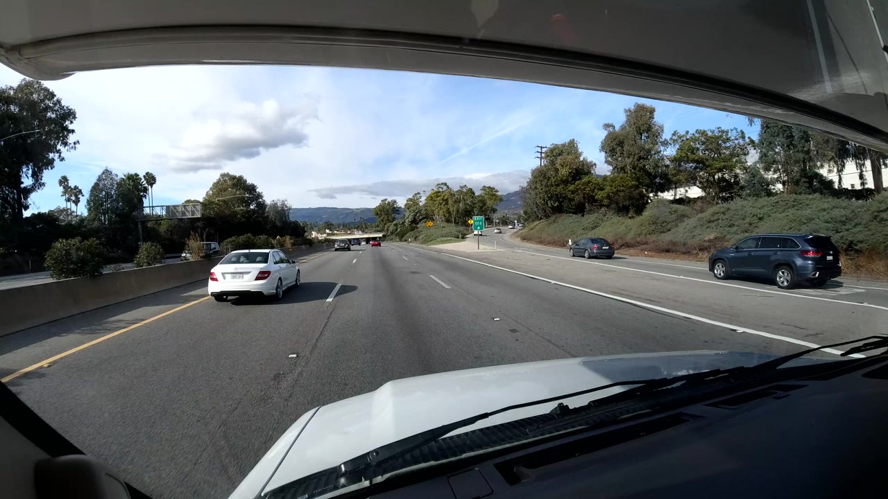 The vehicle follows a white sedan on a Coast California highway, with lane markers, a roadside sign, planted greenery, distant hills, dashboard edges, and no visible people.
