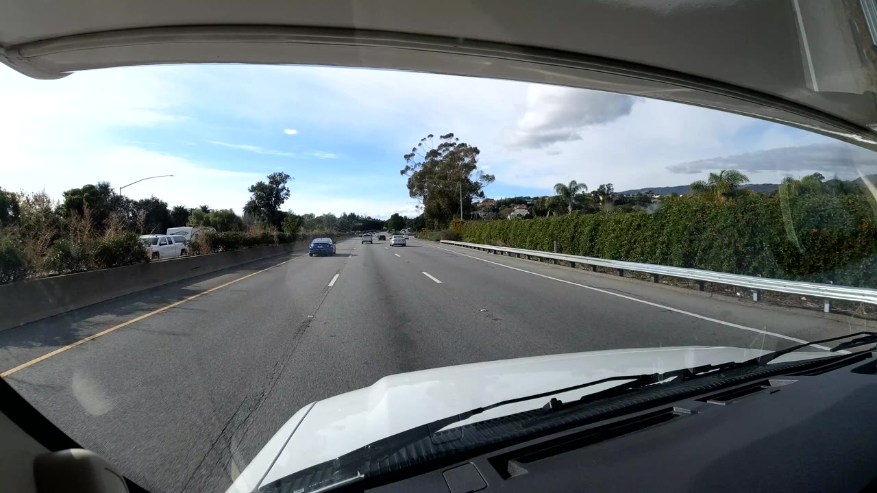 A more open stretch of Coast California highway appears through the windshield, with a dark vehicle near the right side, guardrail, dense roadside greenery, mountains in the distance, and no visible people.