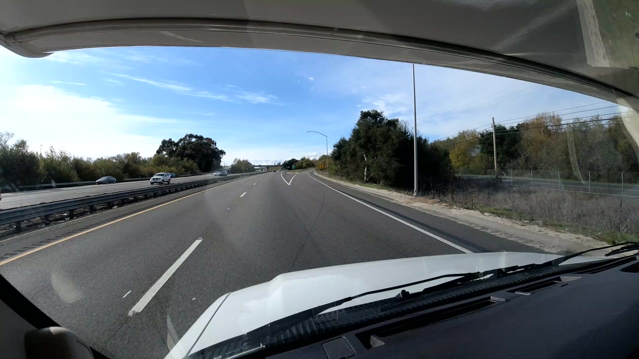 Forward windshield view from a vehicle on a Coast California highway, showing lane markings, roadside trees, a guardrail, and no visible people.