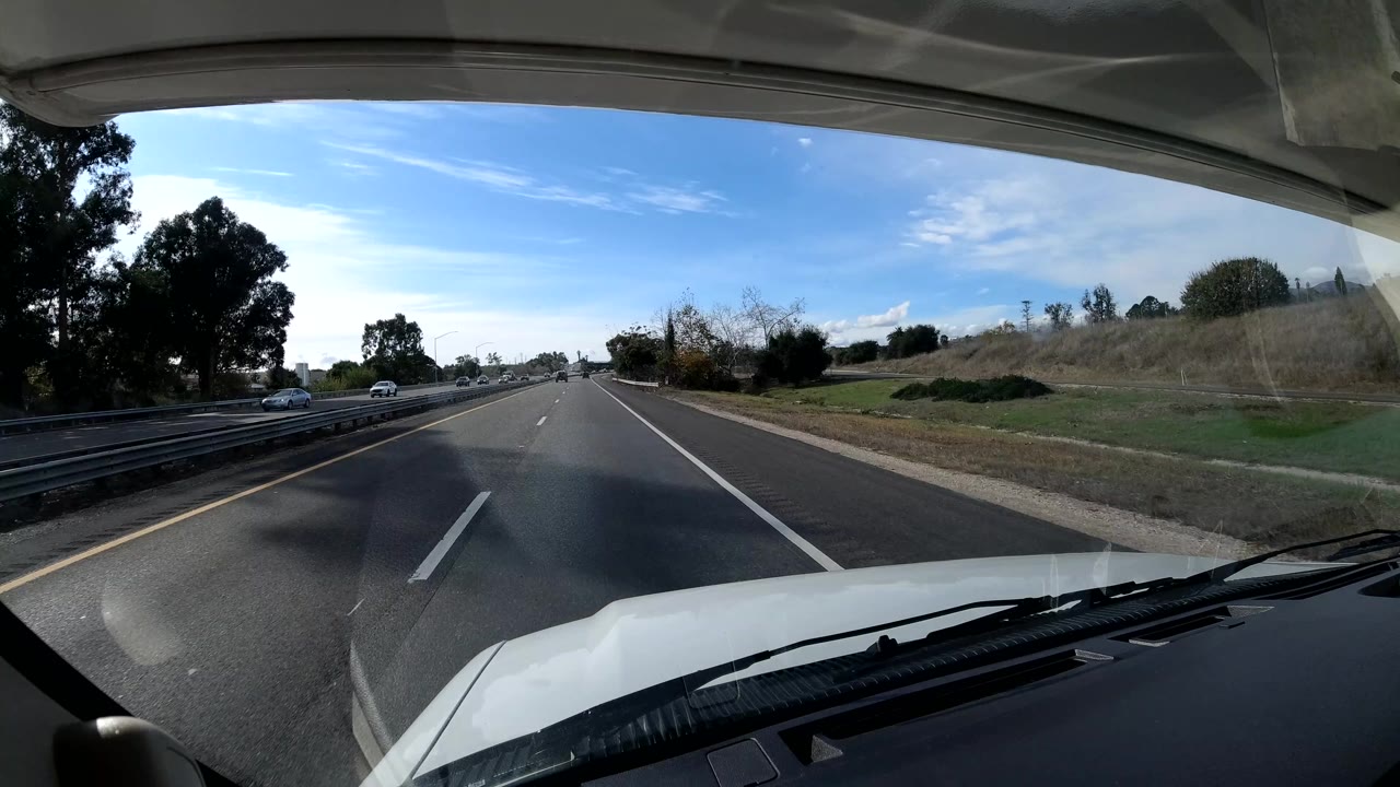 In-vehicle highway perspective in Coast California, with open lanes ahead, roadside brush, trees, a large shadow across the pavement, and no visible people.