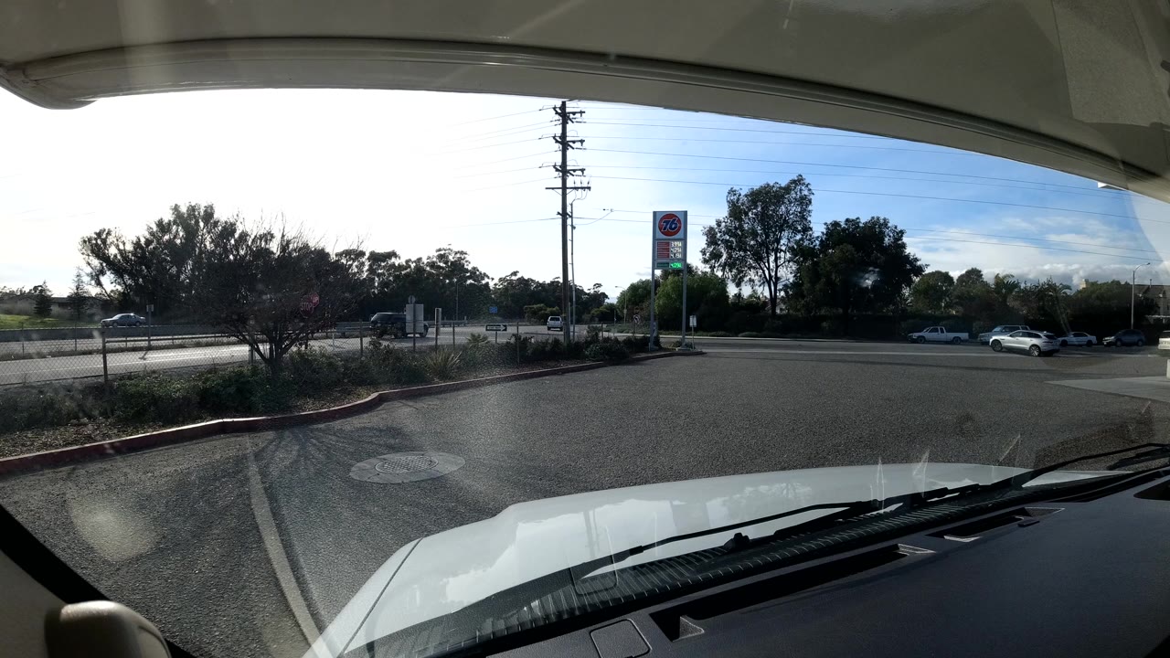 Dash-mounted view in Coast California United States showing the vehicle hood, a roadside lane, trees, utility poles, distant cars, and a tall sign, with no visible people.