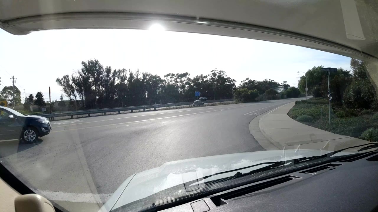Dash view in Coast California United States looking onto a sunlit curved road with a dark car close at right, guardrail, trees, and strong windshield glare, with no visible people.
