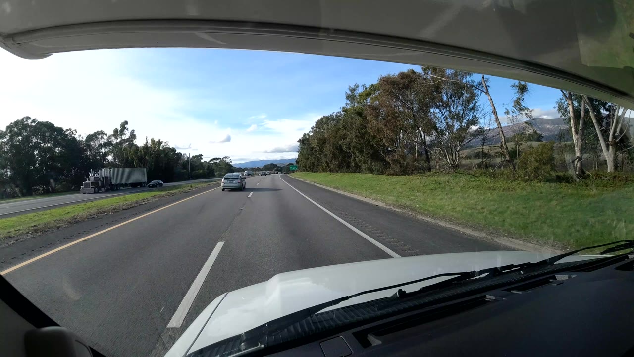 A windshield-framed highway view in Coast, California shows the vehicle hood and wipers at the bottom, cars ahead, roadside trees, grassy shoulders, blue sky, and no visible people.