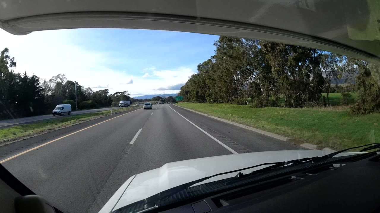 Highway seen through a windshield with a white van on the opposite side and trees ahead.