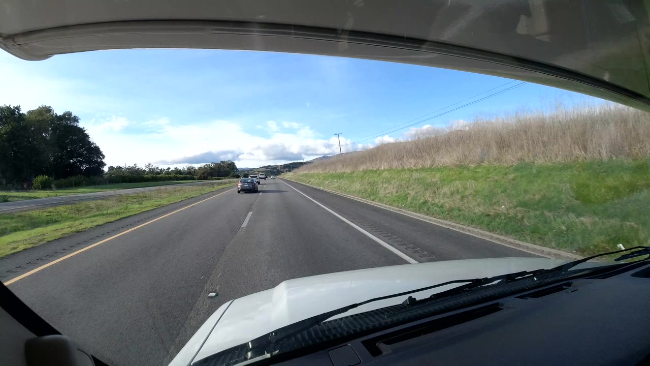 A forward highway view in Coast, California shows a car ahead in the lane, a grassy embankment to the right, trees to the left, utility lines, bright sky, and no visible people.