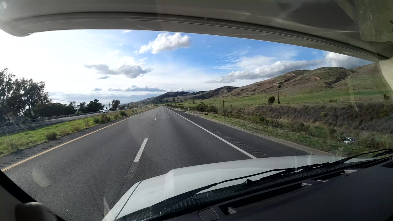 A front-facing vehicle view shows a highway in Coast California, United States, with green hills, roadside brush, dashboard edges, clouds, and no identifiable people visible.