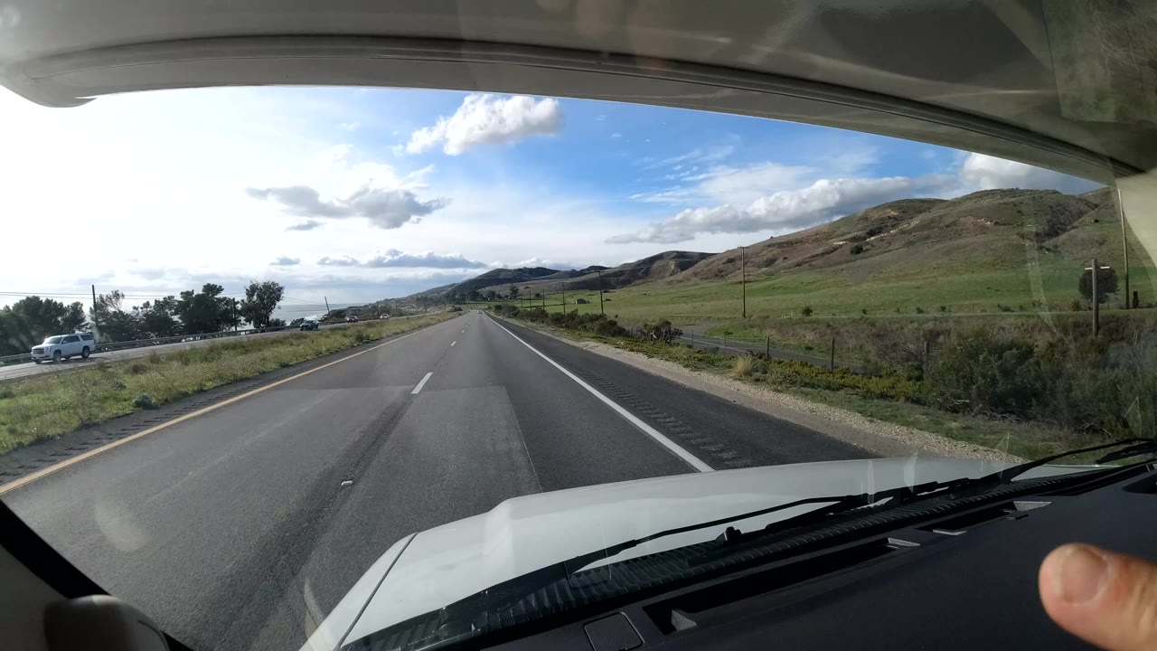 The vehicle continues along a Coast California highway with the dashboard low in frame, hills on both sides, a small car near the left roadside, and no identifiable people visible.