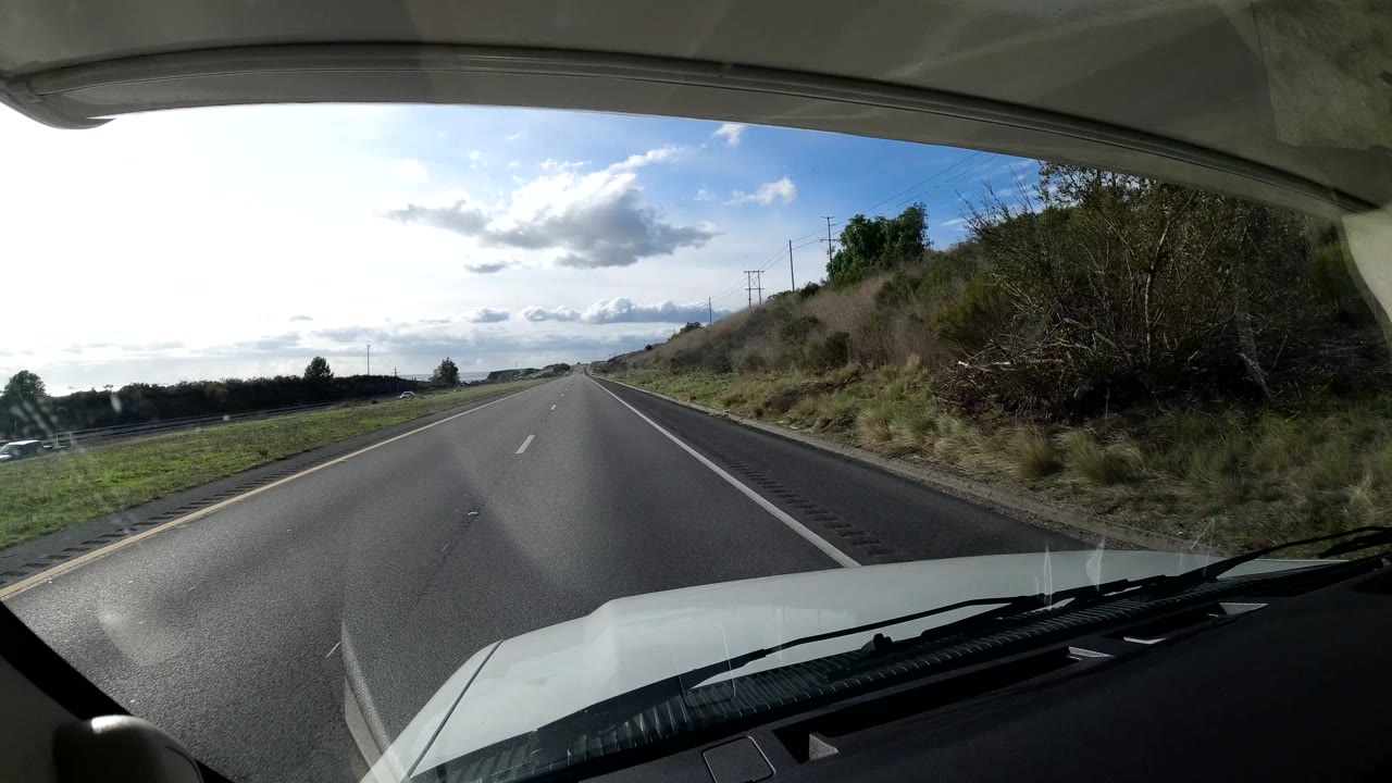 A Coast California road curves slightly ahead beside brush and hillside, with the vehicle hood and windshield visible, a finger entering the lower edge, and no identifiable face visible.
