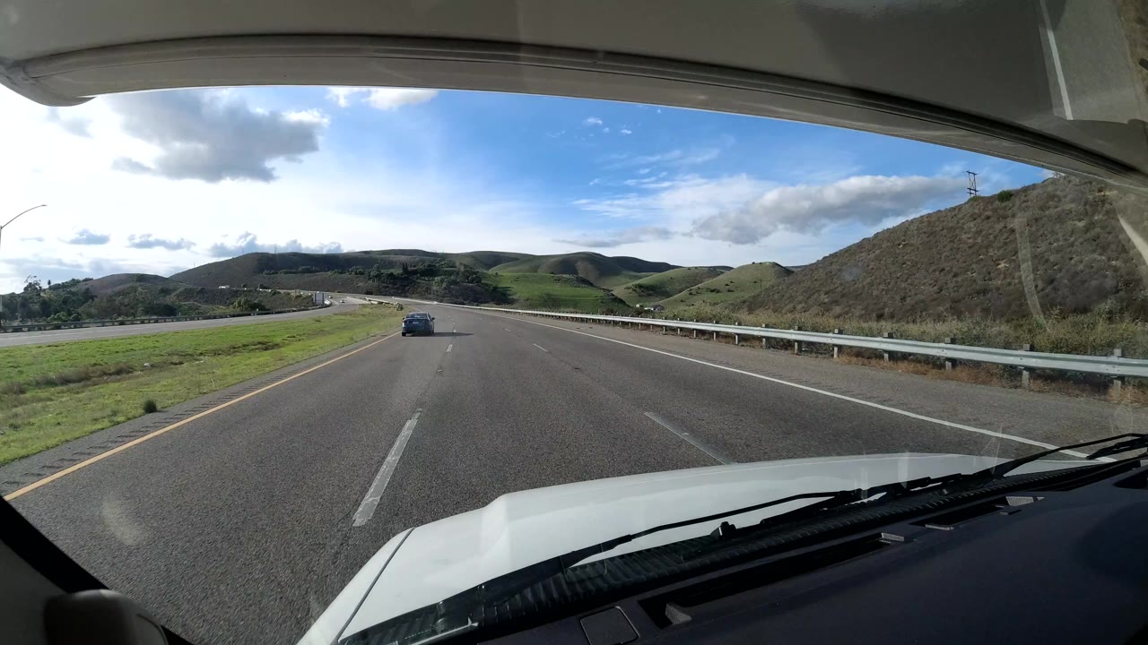 Highway view with guardrail, green median, hills, clouds, and a distant car.