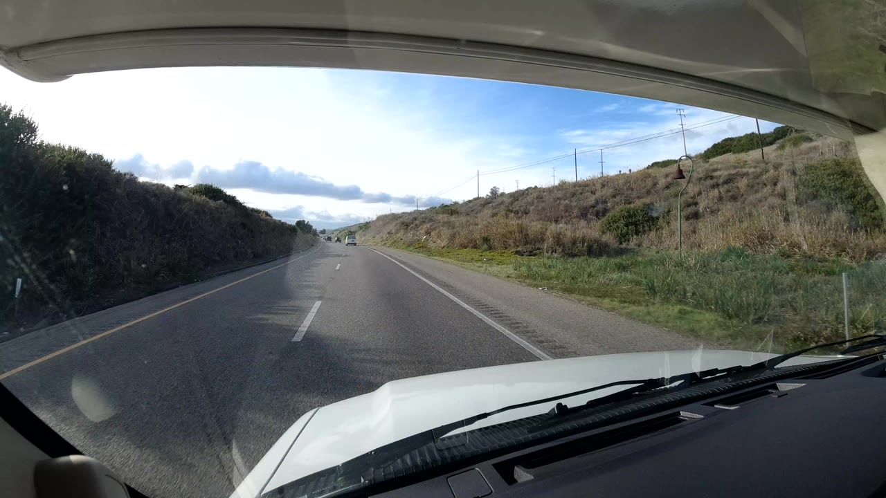 Forward view through a vehicle windshield on a Central Coast California highway, with the hood in the foreground, grassy roadside slopes, distant traffic, cloudy daylight, and no visible people.