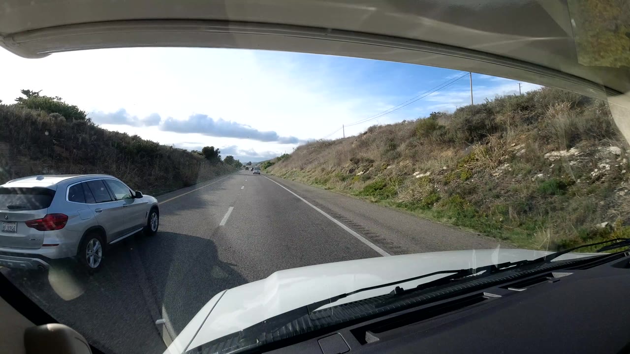A silver SUV travels alongside the camera vehicle on a Central Coast California highway, with the windshield and hood visible, grassy roadside banks, cloudy sky, and no visible people.