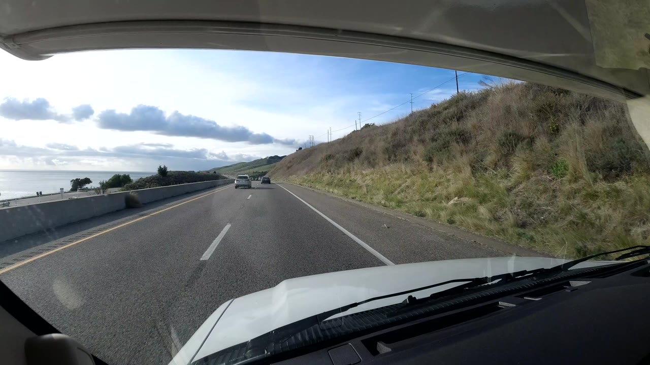 The vehicle approaches cars on a Central Coast California highway with ocean visible beyond a barrier, grassy hillside on the right, cloudy daylight, and no visible people.