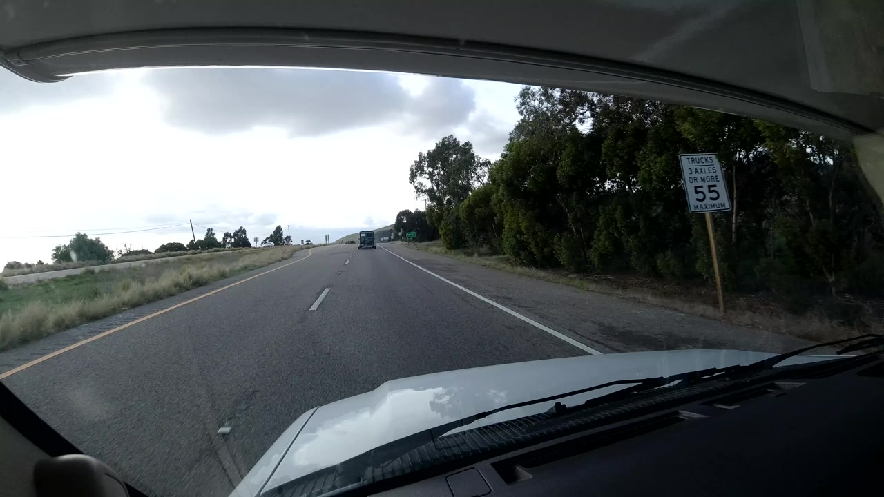 Windshield view from a vehicle on a Coast California highway, with the hood and wipers in the foreground, a 55 mph speed sign on the right, trees along the shoulder, and no visible people.