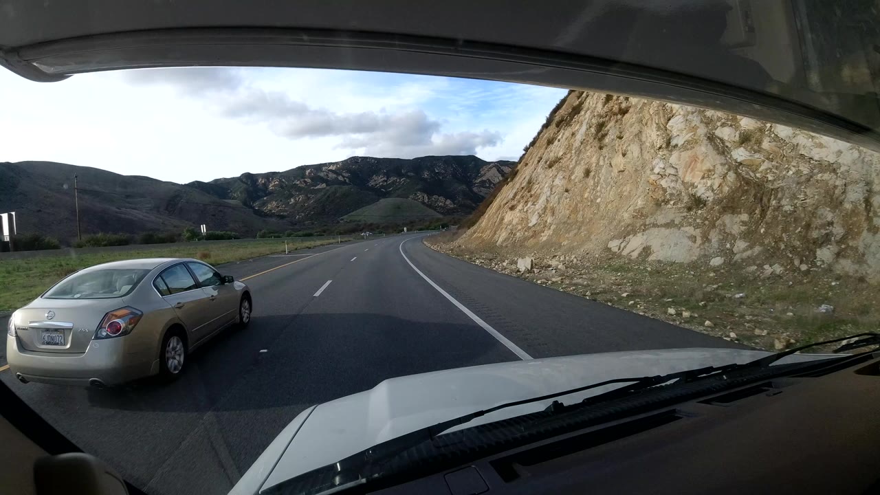 The windshield view shifts to a curving Coast California roadway beside a rock cut, with a silver sedan nearby, mountain slopes beyond the lanes, and no visible people.
