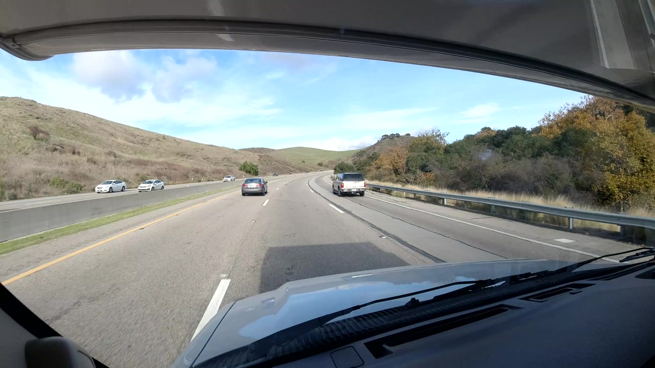 Vehicle windshield view of highway traffic with hills and shrubs along the shoulder.
