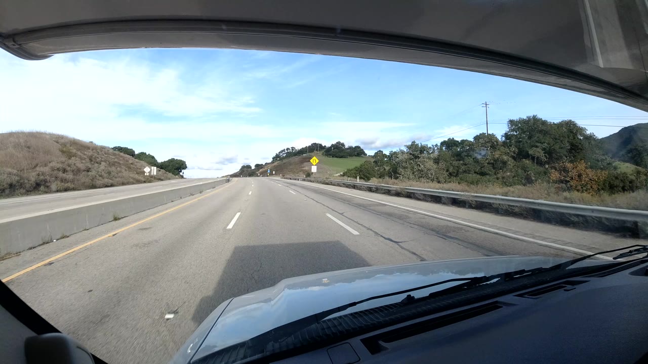 A more open stretch of Central Coast California highway appears through the windshield, with fewer nearby cars, guardrails, roadside signs, hillside vegetation, and no visible people.