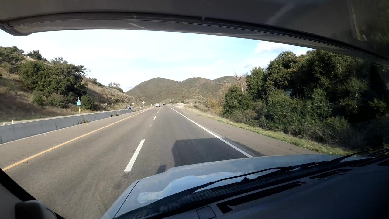 A forward windshield view on the California coast shows the vehicle hood, open divided highway lanes, green roadside trees, low hills, and no visible people.