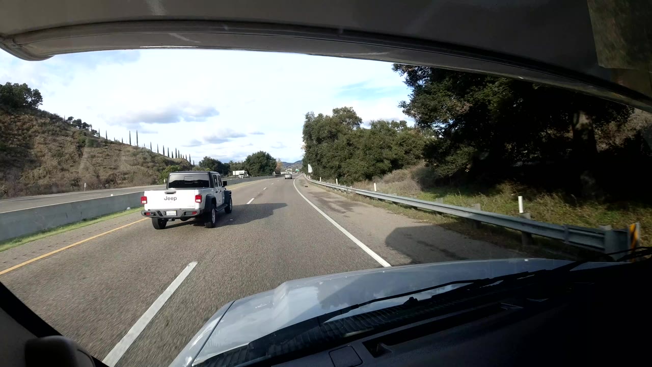 A white pickup drives ahead on the California coast highway, with guardrails, trees, hillside vegetation, the vehicle hood, and no visible people in frame.