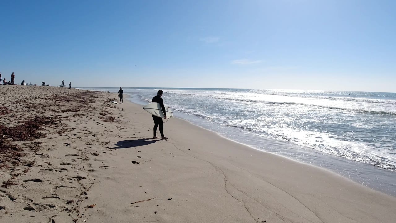 Surfer with white board walking beside reflective ocean water.