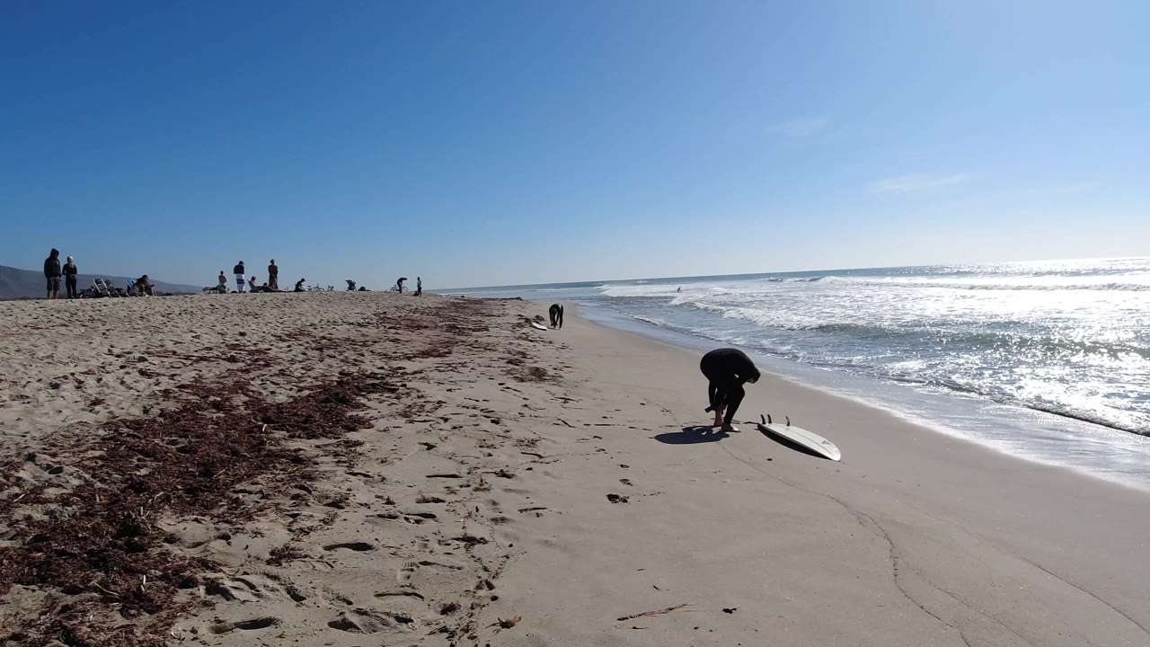 Beach shoreline with bright waves, distant people, and a surfboard near the water.