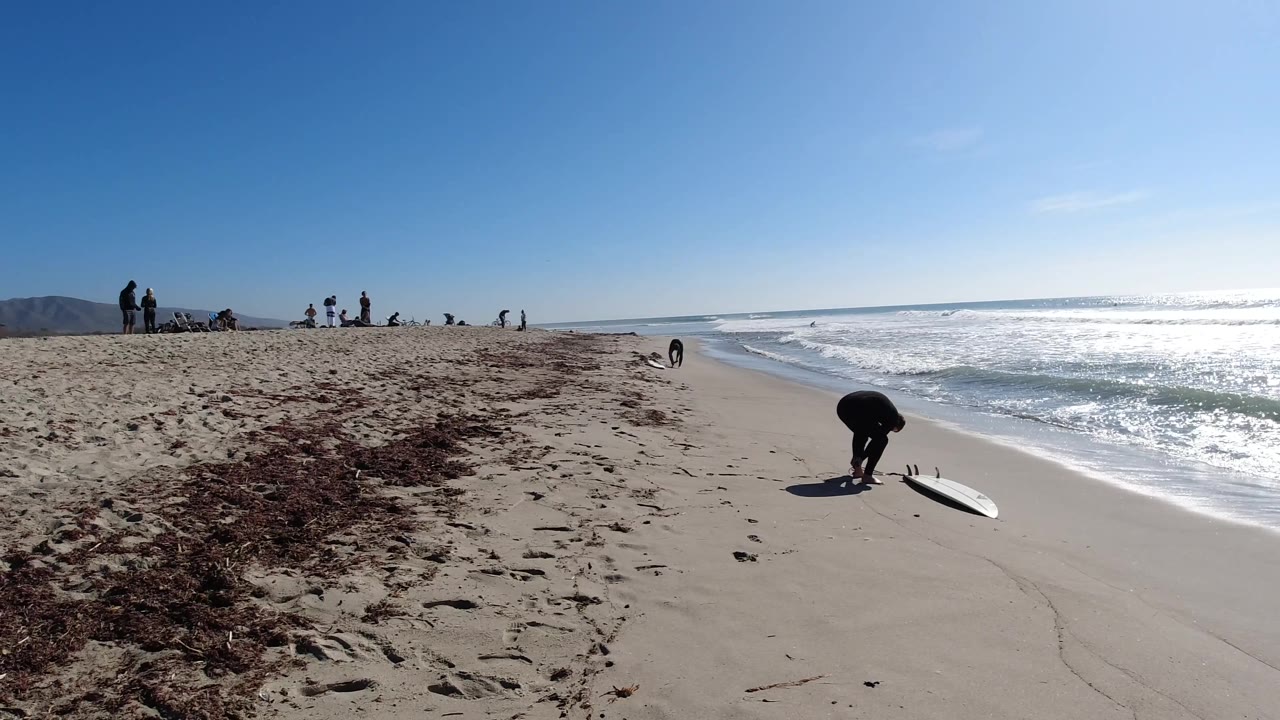At Trestles Beach, California, the frame shows a wide sandy foreground, seaweed patches to the left, scattered beachgoers near the horizon, and surf breaking to the right.