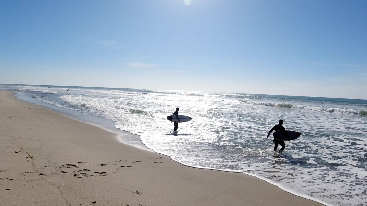 At Trestles Beach, California, two surfers in dark wetsuits carry surfboards from wet sand toward bright foamy waves under a clear blue sky.