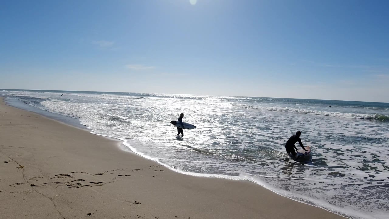At Trestles Beach, California, the surfers have moved farther into the shallow break, with one near the center carrying a board and the other bending near the foam.