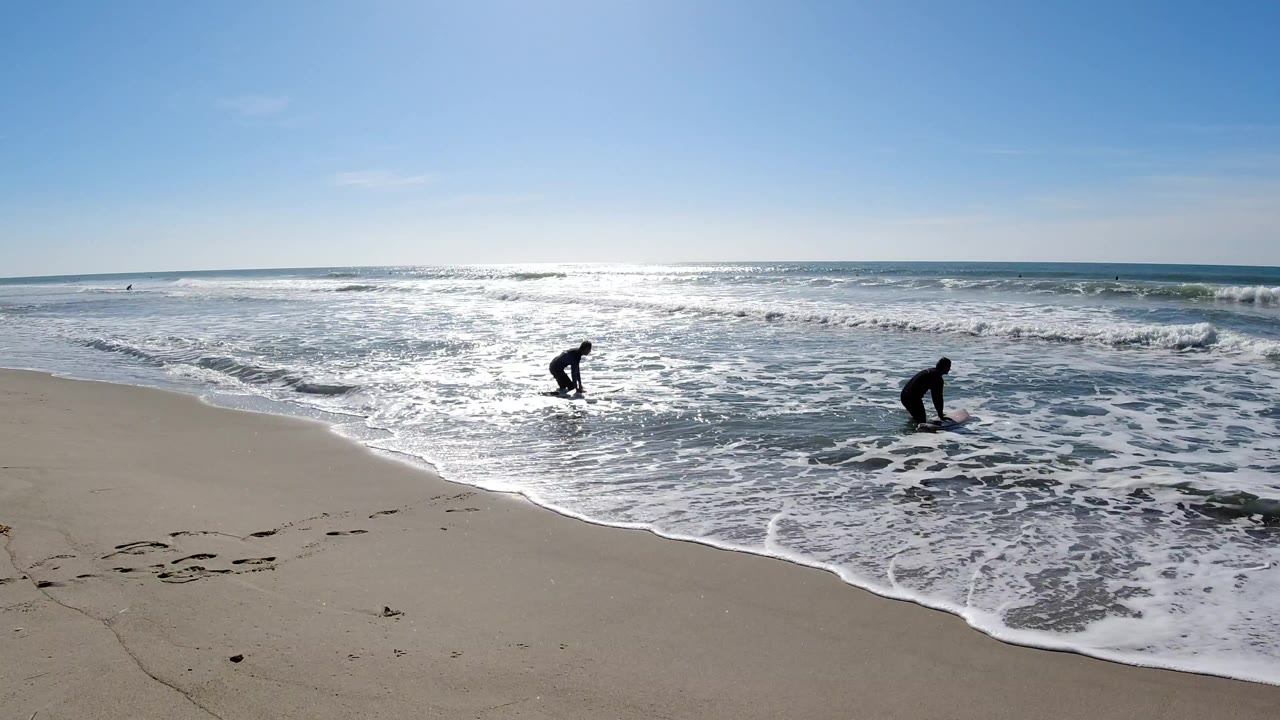At Trestles Beach, two wetsuit surfers stand near the shallow whitewater as bright sun reflects across the ocean and wet sand.