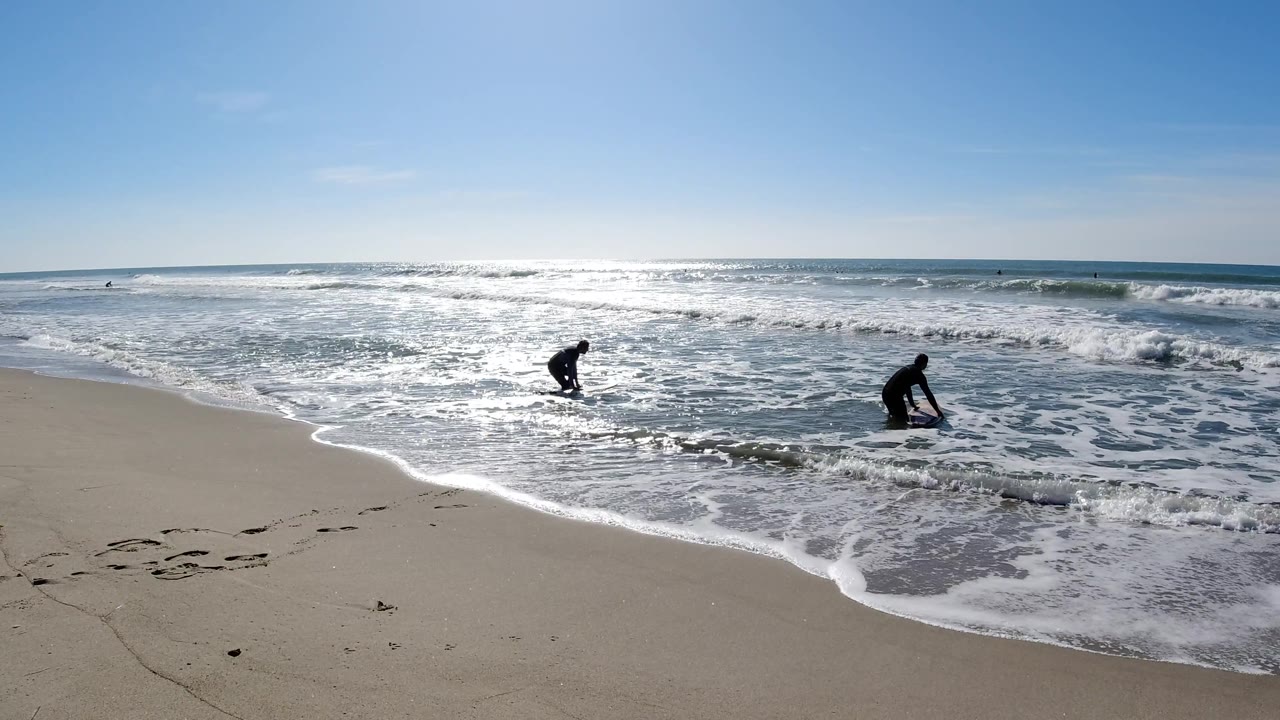 At Trestles Beach, the right surfer is lower in the wash with a board while the left surfer remains near a band of whitewater under a clear sky.