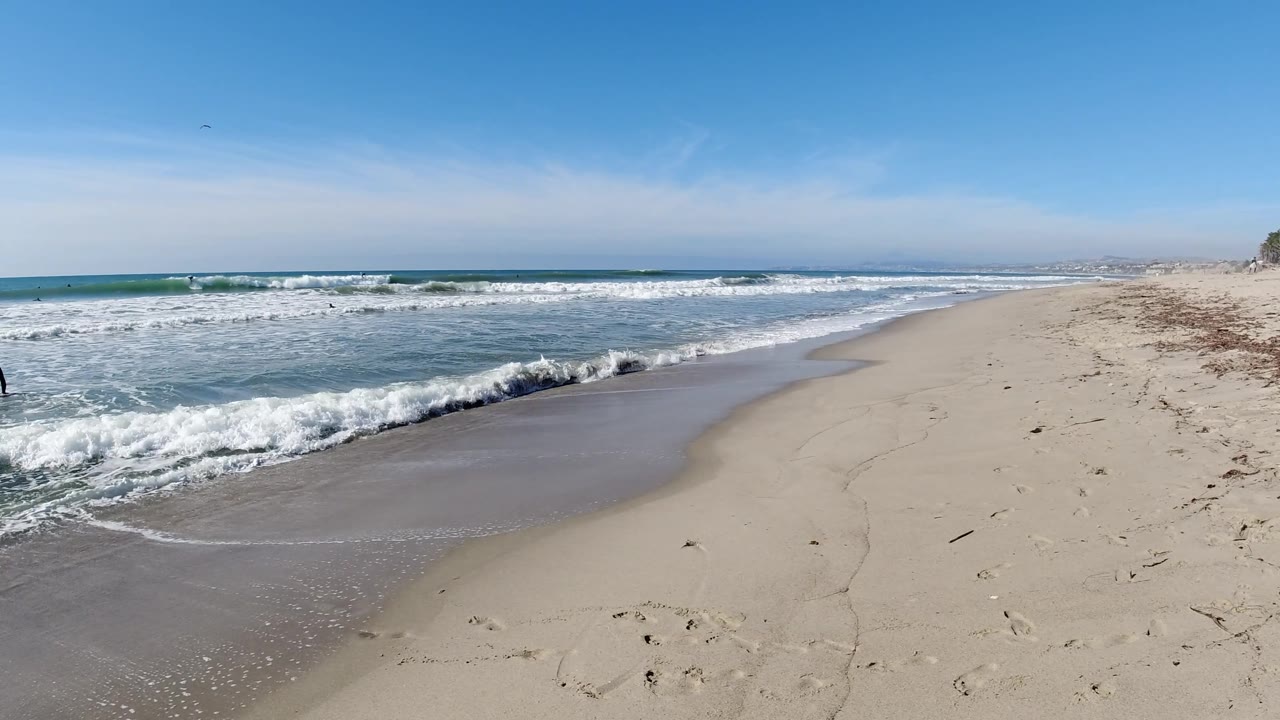 A small distant bather appears near the water at Trestles Beach, California as white waves break across the left side and the sandy beach opens to the right.
