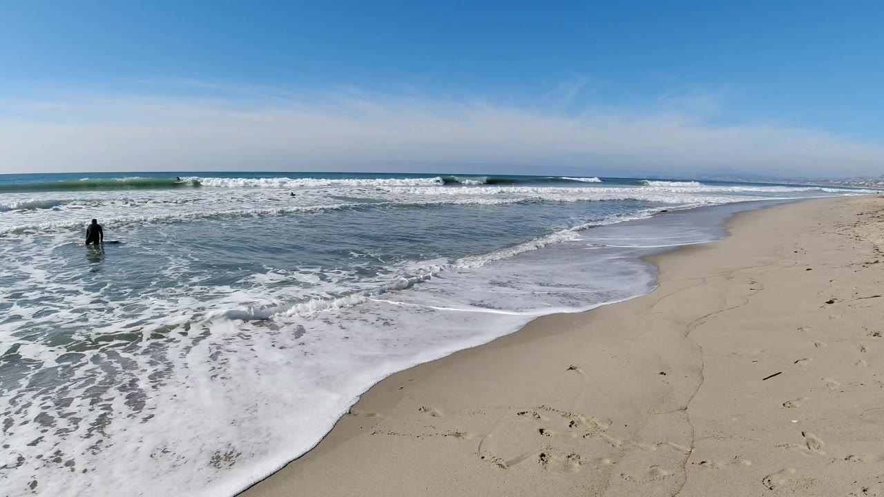 At Trestles Beach, California, the tide spreads over wet sand while one distant person stands near the left edge of the surf and white waves form behind them.