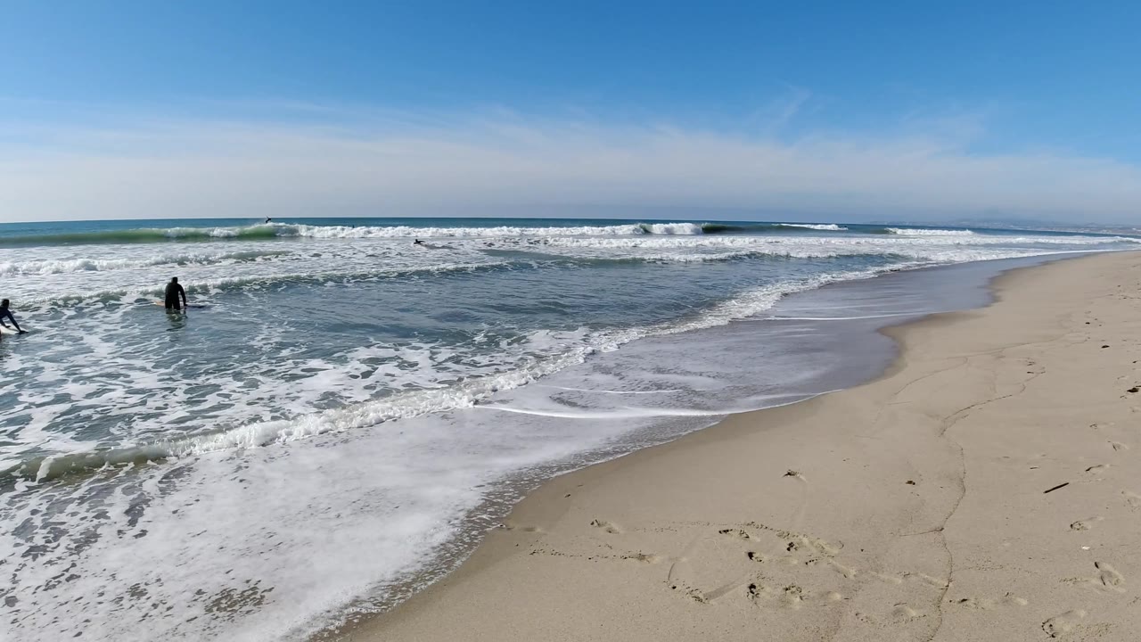At Trestles Beach, California, two people are visible in the surf zone, one lower to the water and one upright, with a breaking wave and open sand to the right.