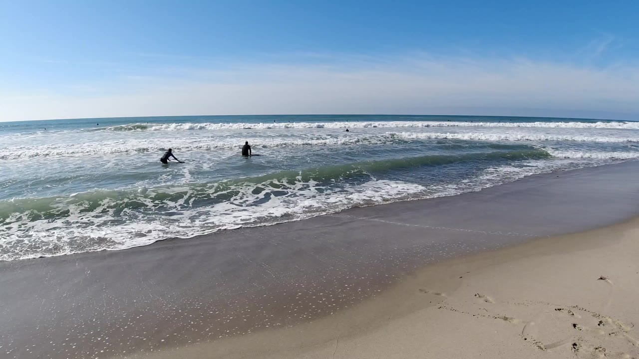 Wide elevated view of Trestles Beach, California, showing two small dark figures entering the surf zone amid rolling green-blue waves and white foam wash, with wet sand in the foreground and a pale blue sky above; people are visible as small distant silhouettes.