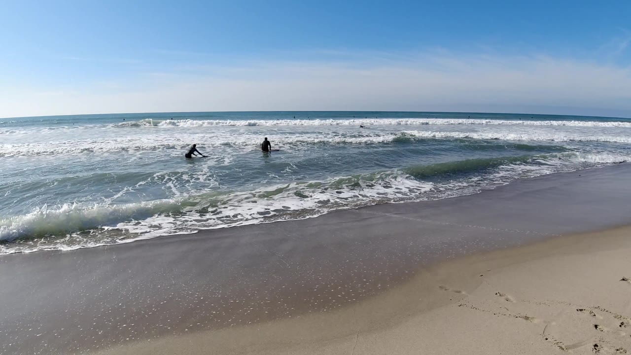 Wide elevated view of Trestles Beach, California: two small dark figures in the surf zone as another wave crests and white foam spreads across the shoreline, wet sand in the lower foreground, pale blue sky above; people are visible as small distant silhouettes.