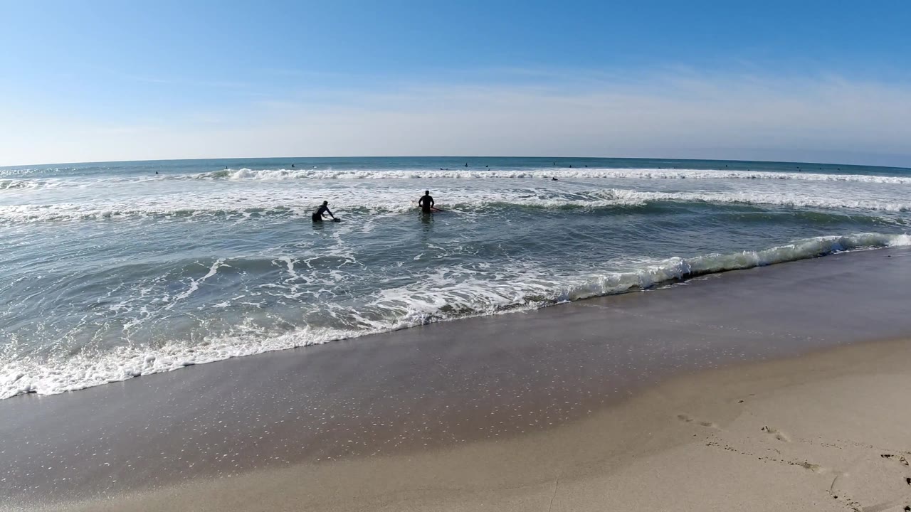 Elevated beach view with prominent white foam wave wash across the sand, two small dark figures in the surf, and pale blue sky.