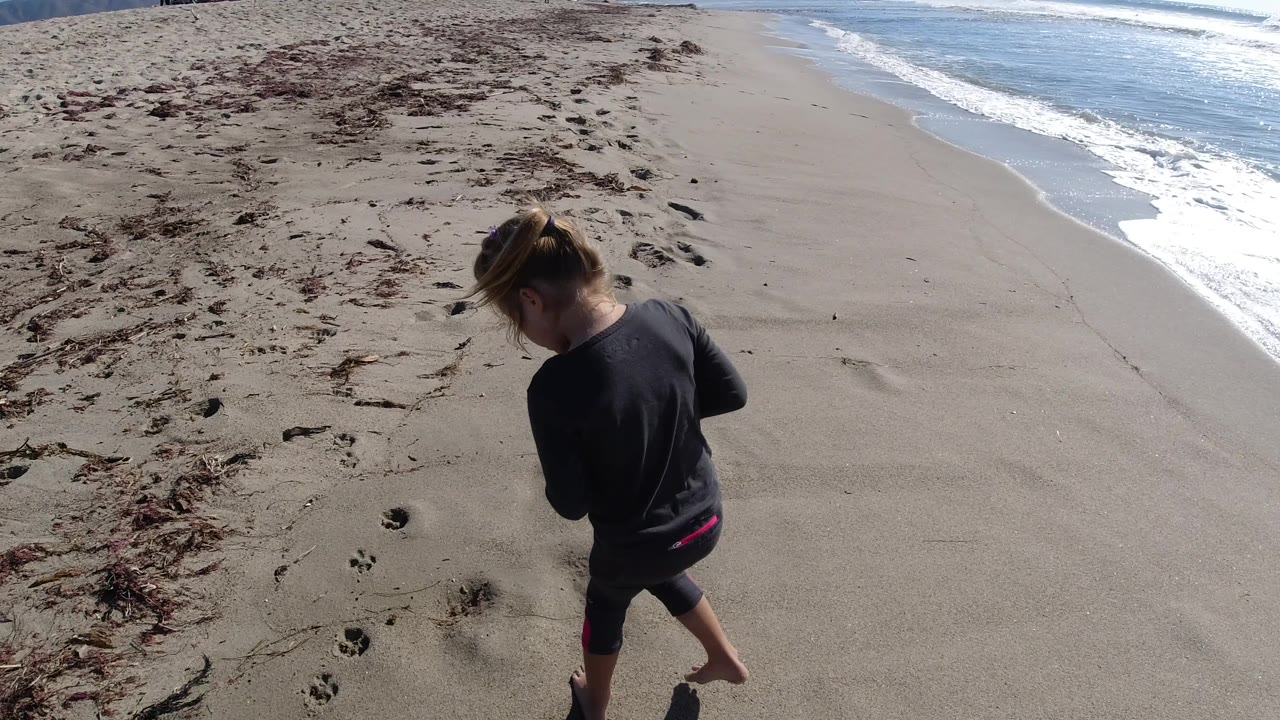 At Trestles Beach, California, the girl remains near the lower left of the frame with scattered seaweed and a diagonal band of surf behind her.