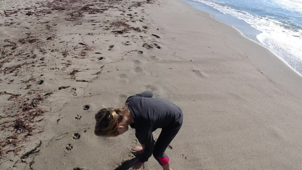 At Trestles Beach, California, the girl bends toward the sand near footprints and seaweed while the ocean edge stays visible at upper right.