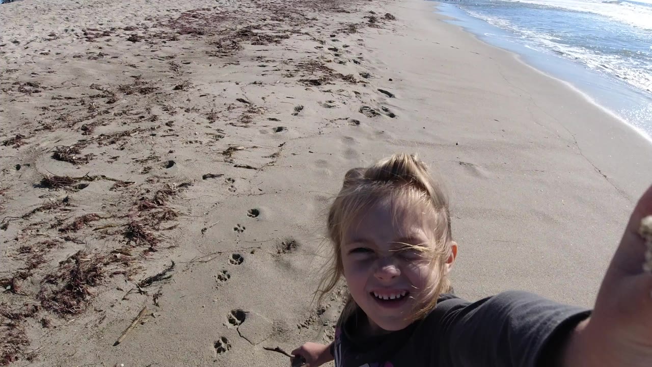 A smiling blonde young girl is seen close to the camera at Trestles Beach, California, with pale sand, seaweed patches, footprints, and ocean foam behind her.