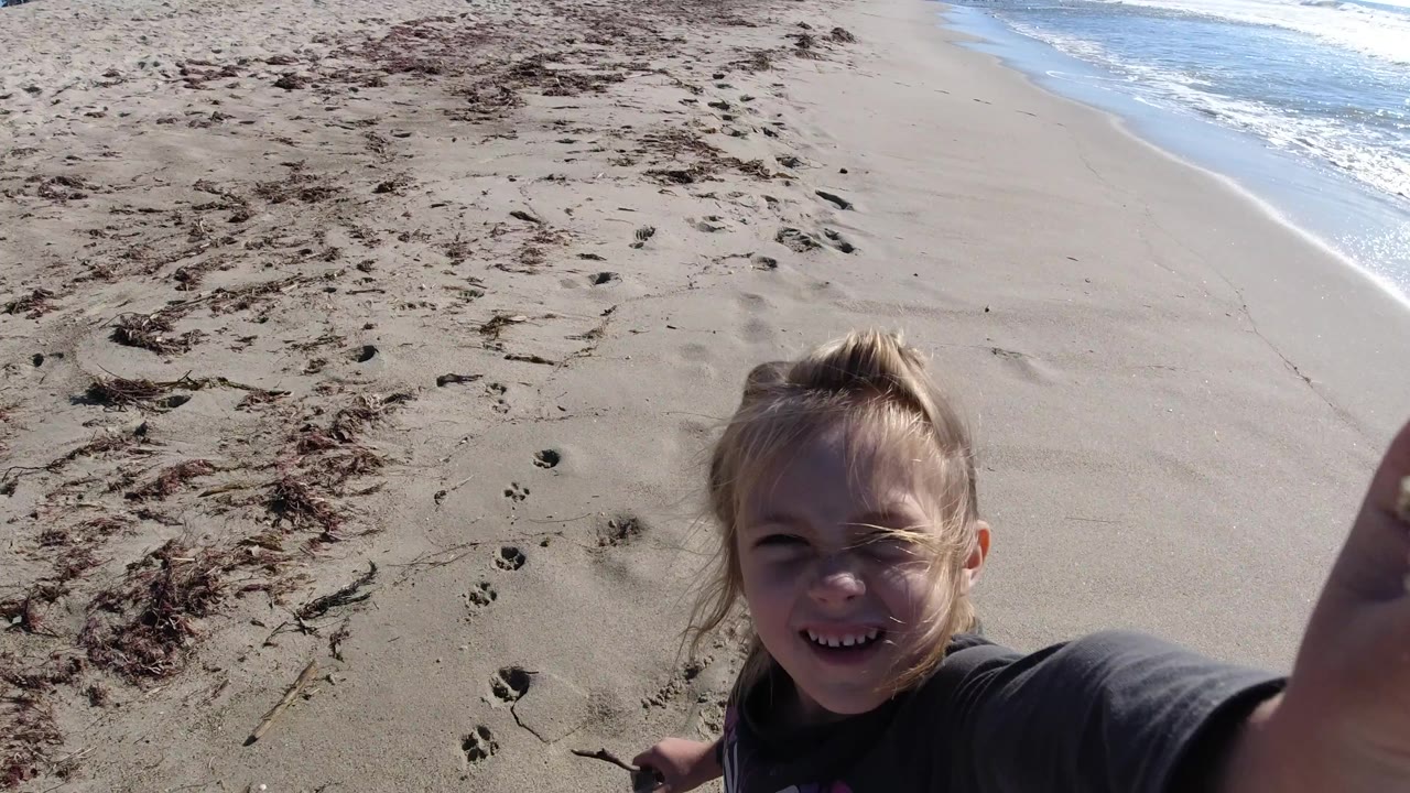 The young girl remains close to the lens at Trestles Beach, California, smiling in bright sun as the sandy shoreline and white surf angle across the background.
