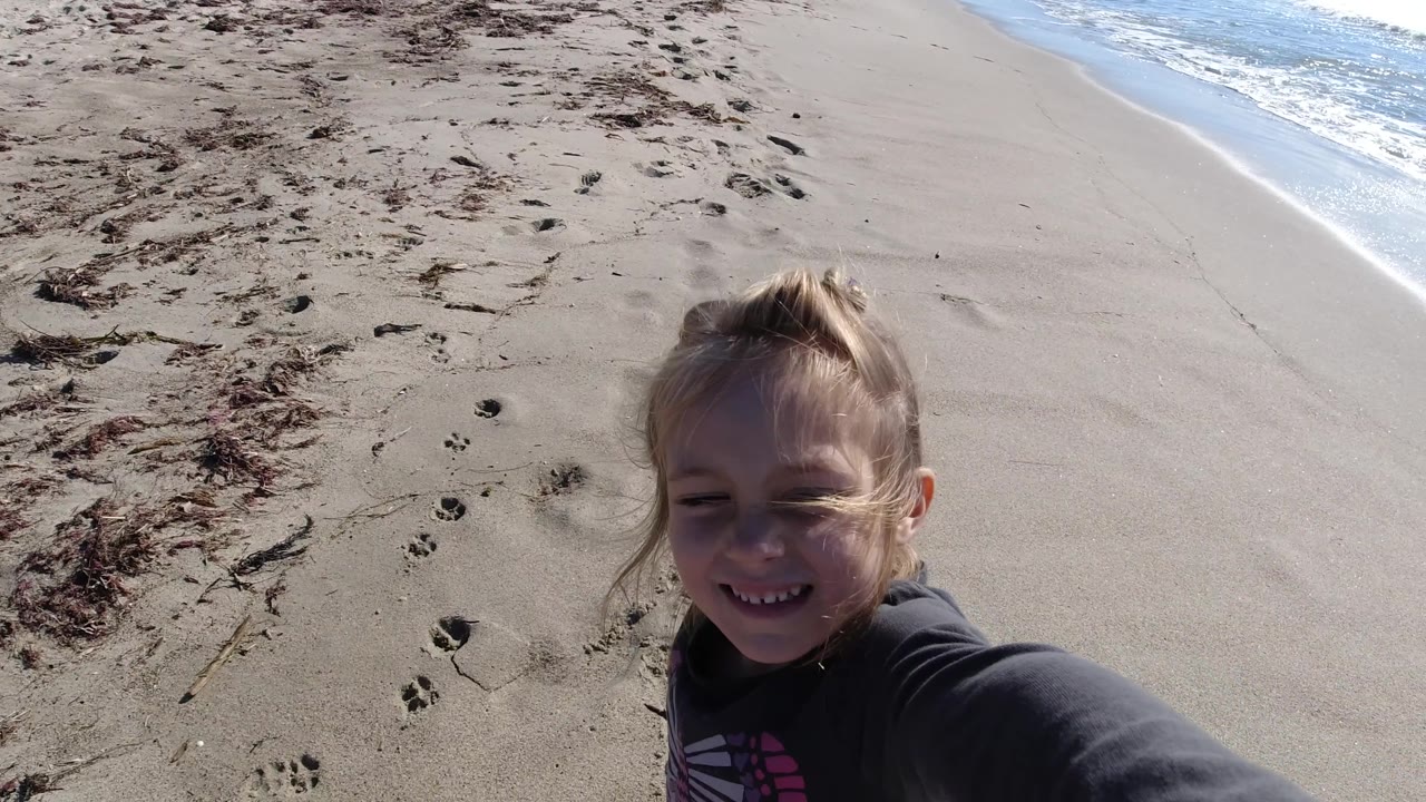 A close high-angle view shows the young girl smiling beside the sandy edge of the surf at Trestles Beach, California, with windblown hair and blue water behind her.