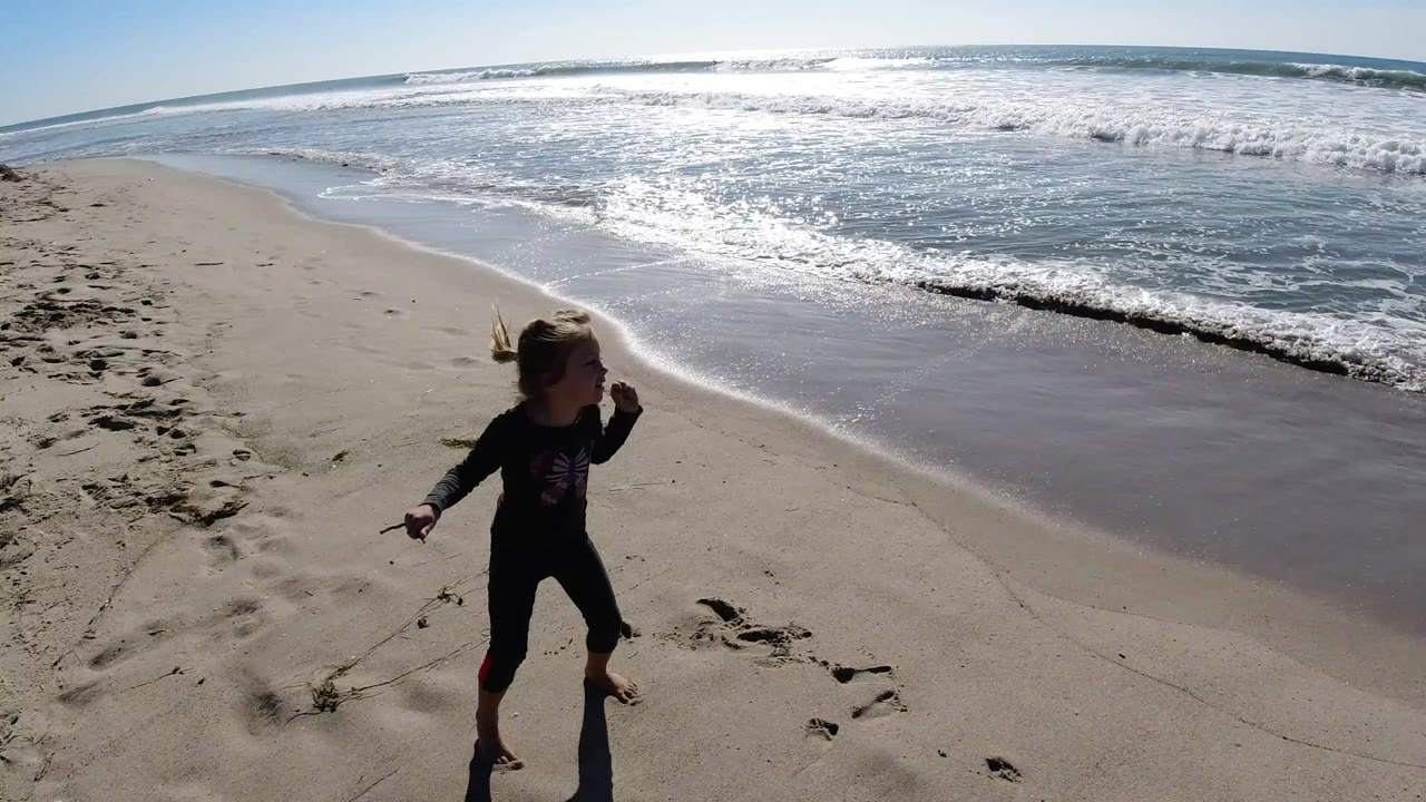 At Trestles Beach, California, a little girl in dark beachwear stands near footprints on dry sand while bright white surf rolls in behind her.