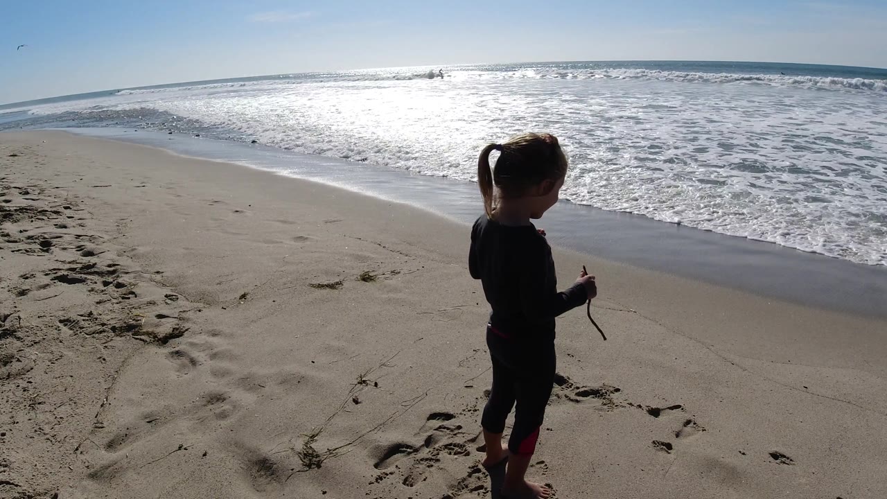 A young girl stands at the right edge of the beach frame holding a stick, with foamy water and a bright ocean horizon stretching behind her at Trestles Beach, California.