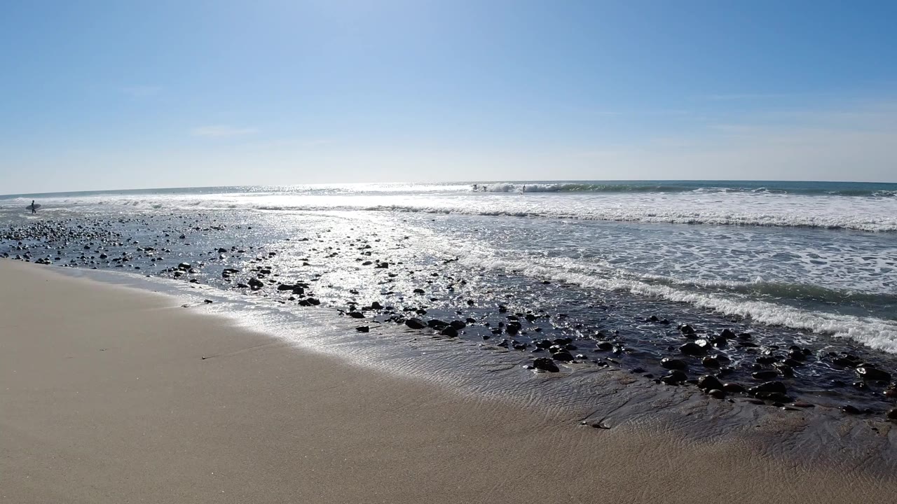 Wide view of Trestles Beach, California, with bright ocean glare, scattered rocks on wet sand, breaking waves, and no visible people.