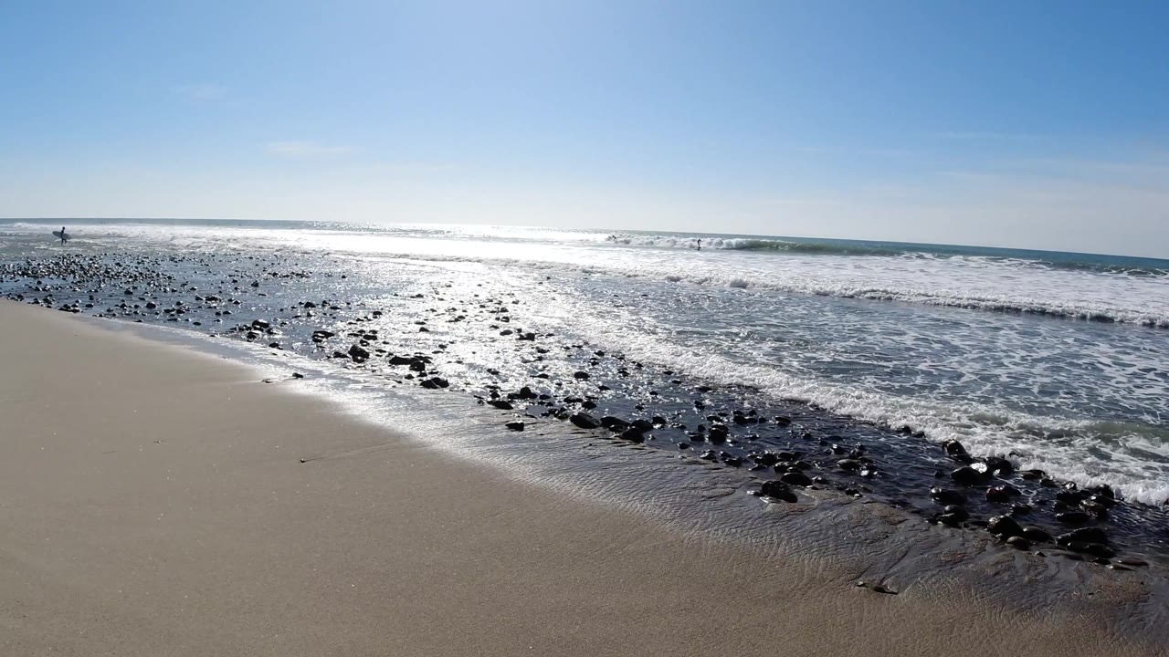 Bright surf at Trestles Beach breaks behind a diagonal band of dark rocks on wet sand, with strong glare on the water and no visible people.