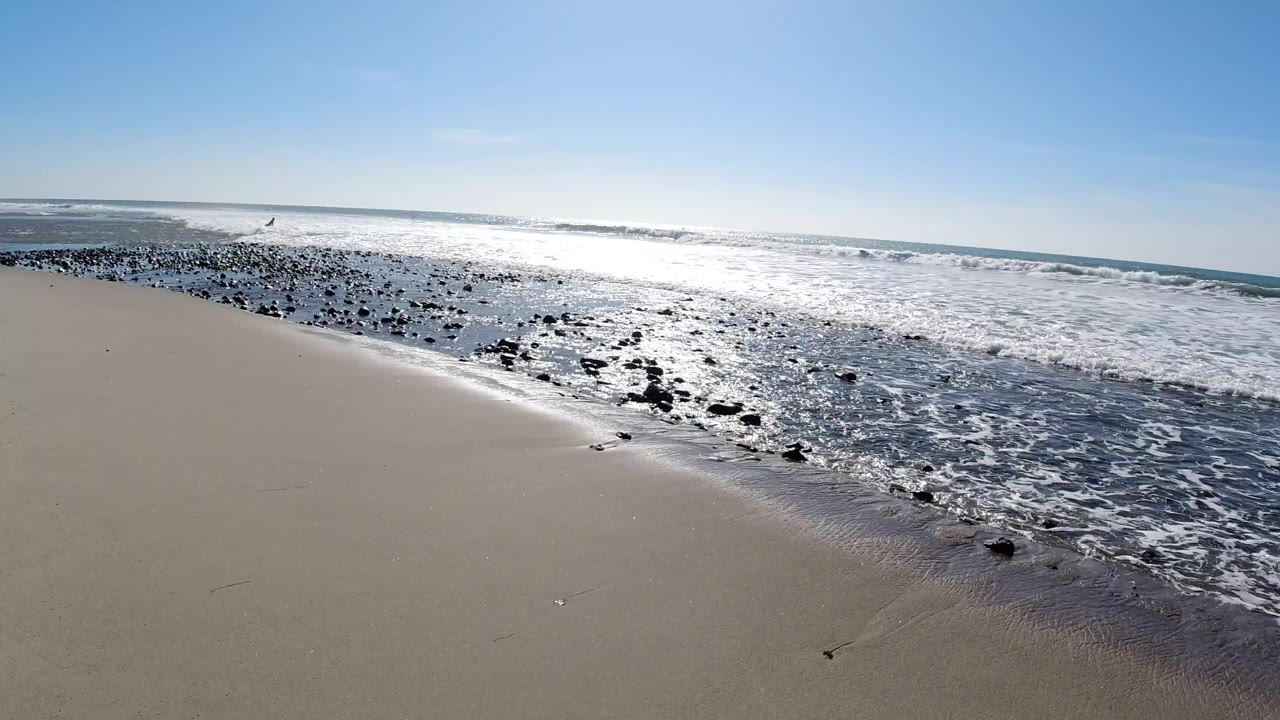 Trestles Beach, California, opening frame: pale sand dominates the foreground, dark rocks line the waterline with white foam surf, the ocean glitters under direct sun, and the sky is clear blue; no visible people.