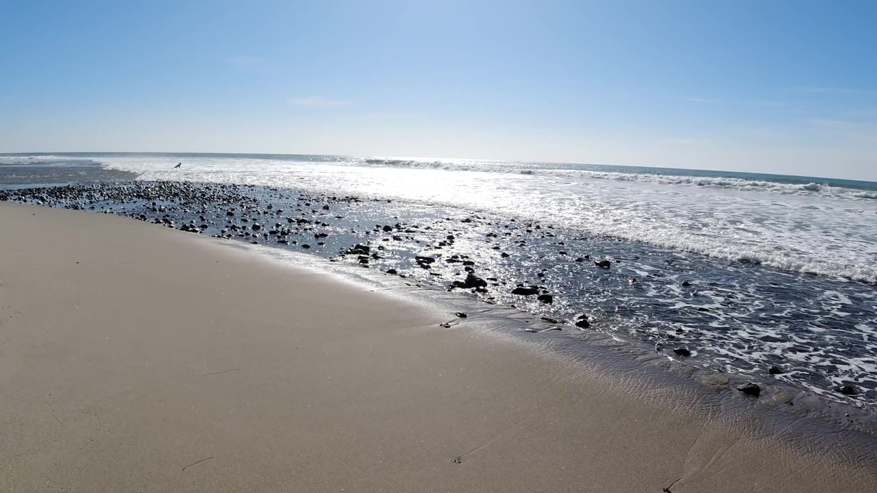 Trestles Beach, California, third frame: pale sand foreground, a band of dark rocks curving along the shoreline with white surf, glittering ocean, and blue sky; no visible people.