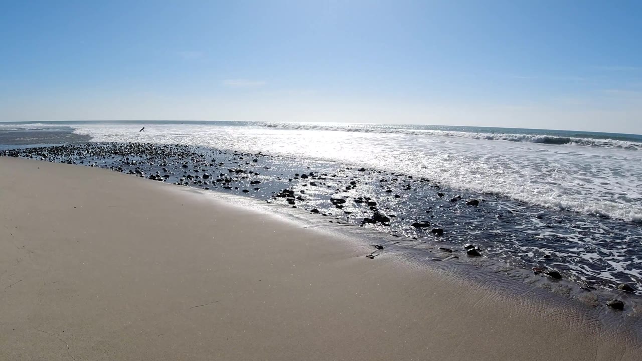 Beach with sand foreground, dark rocks and white surf at the waterline, ocean horizon, blue sky, and small dark specks near the water that may be birds.