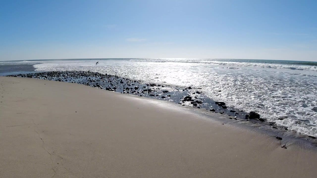 Wide pale sandy beach at Trestles Beach, California, with white wave foam and dark wet rocks curving diagonally across the midground, bright sun glare on the ocean surface, and no visible people